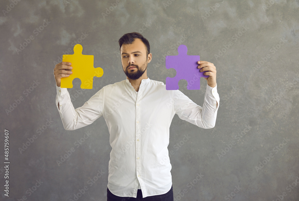 Portrait of serious pensive guy standing against grey studio background joining two jigsaw puzzle pieces of yellow and purple color as metaphor for finding good professional solution. Business concept Portrait of serious pensive guy standing against grey studio background joining two jigsaw puzzle pieces of yellow and purple color as metaphor for finding good professional solution. Business concept