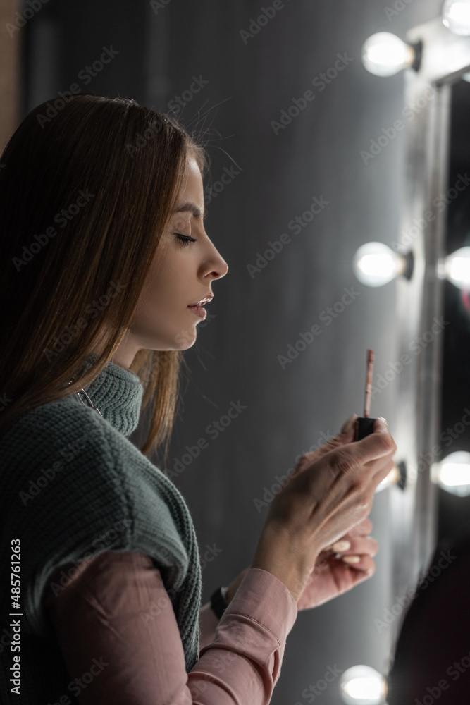 Obraz premium beautiful young girl in fashionable clothes does makeup and paints her lips near a vintage mirror with lights bulbs in the dark