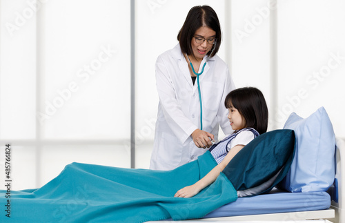 Asian professional friendly female doctor in white lab coat and stethoscope standing smiling to young girl patient lay down on bed in hospital wardroom photo