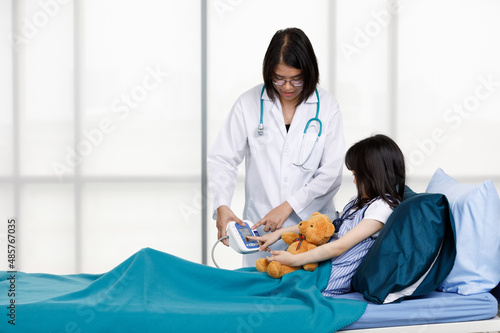 Asian professional friendly female doctor in white lab coat and stethoscope standing smiling to young girl patient lay down on bed hugging teddy bear doll in hospital wardroom photo