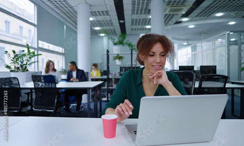 Black business woman smiling while communicating online via laptop in open space office, diverse coworkers on background. Cheerful manager in coworking. Concept of business