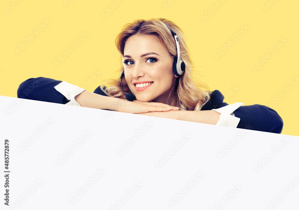 Call center. Customer support service female phone operator or sales agent in headset, confident suit standing behind signboard with copy space area for text, isolated over yellow background Call center. Customer support service female phone operator or sales agent in headset, confident suit standing behind signboard with copy space area for text, isolated over yellow background