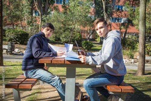 Two young university students studying in a university garden with a laptop computer and papers looking at the camera.