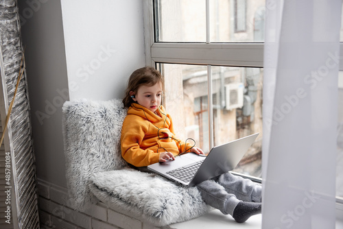 Little toddler girl sits on wide windowsill, plays games on laptop, listens to music on headphones