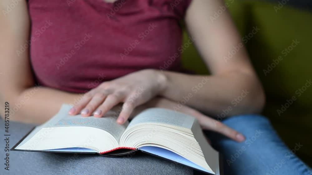 Selective focus on book read by caucasian woman with short haircut in burgundy T-shirt sitting on gray sofa. Concept of time alone with yourself. Selective focus on book read by caucasian woman with short haircut in burgundy T-shirt sitting on gray sofa. Concept of time alone with yourself.