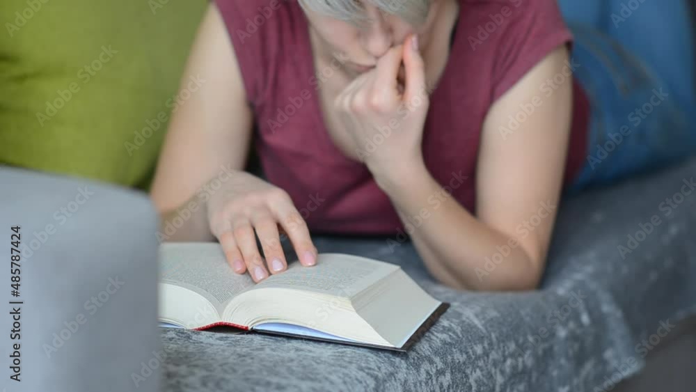 Selective focus on book read by caucasian woman with short haircut in burgundy T-shirt and jeans lying on gray sofa with pillows. Concept of time alone with yourself. Selective focus on book read by caucasian woman with short haircut in burgundy T-shirt and jeans lying on gray sofa with pillows. Concept of time alone with yourself.