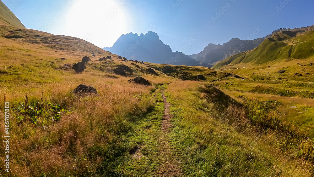 Fototapeta premium A panoramic view on the sharp mountain peaks of the Chaukhi massif in the Greater Caucasus Mountain Range in Georgia, Kazbegi Region. A hiking trail on a green alpine pasture. Georgian Dolomites.
