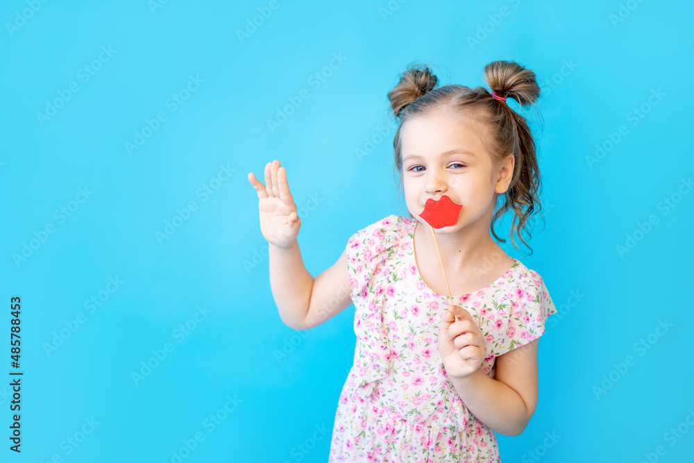 a little child girl on a blue isolated studio background with a paper accessory on a stick with her lips is having fun and fooling around. Space for copying text. Cute kid a little child girl on a blue isolated studio background with a paper accessory on a stick with her lips is having fun and fooling around. Space for copying text. Cute kid