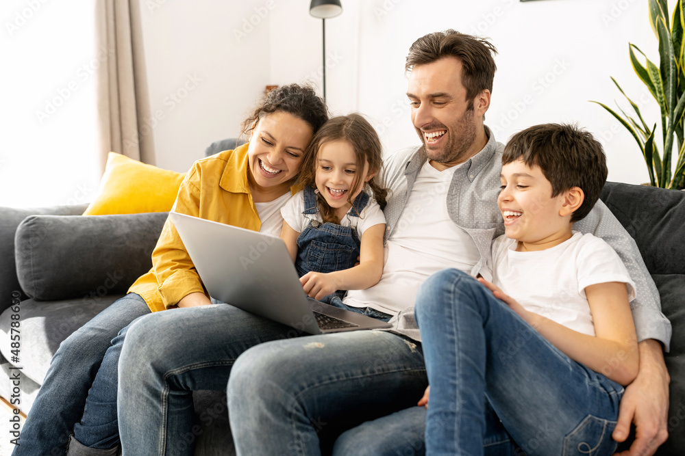 Cheerful happy family of four using laptop sitting on the sofa in cozy apartment, parents and two kids watching movie on computer, comedy, spends time on the weekend together at home Cheerful happy family of four using laptop sitting on the sofa in cozy apartment, parents and two kids watching movie on computer, comedy, spends time on the weekend together at home