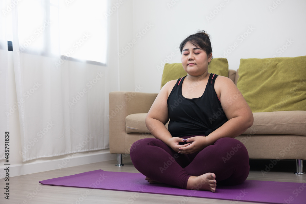 Young asian sport woman plus size sitting on the yoga mat practicing  meditation. Fitness or exercise at home. Beautiful female relaxing after  workout. Stock Photo