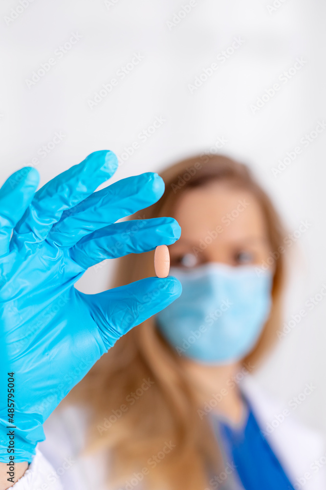 Young female doctor with medical mask and blue gloves holding a pill, tablet. Medicine, healthcare during Covid-19, pharmaceuticals. Doctor with white coat, ready to help. Copy space. Vertical view. Young female doctor with medical mask and blue gloves holding a pill, tablet. Medicine, healthcare during Covid-19, pharmaceuticals. Doctor with white coat, ready to help. Copy space. Vertical view.