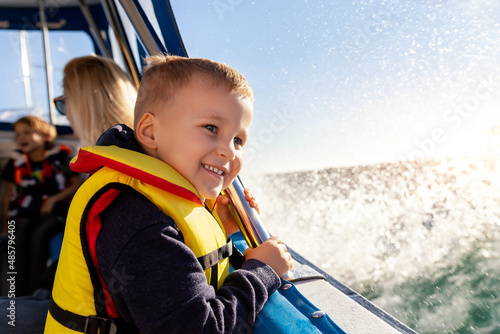 Portrait of cite little blond happy excited smiling caucasian boy wear lifevest enjoy sailing on motor boat sea against blue sky and water splash wave sun backlit. Summer travel vacation recreation