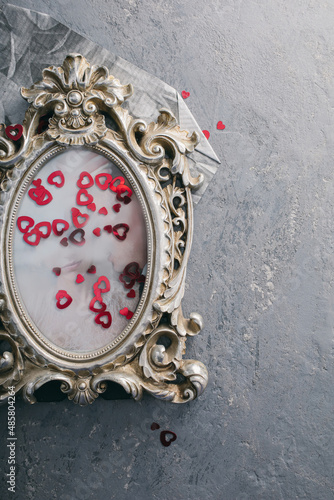 A concrete gray background with a vintage silver photo frame, in which a photo of young people in love is inserted, next to it lies a scattering of red sequins in the shape of a heart.