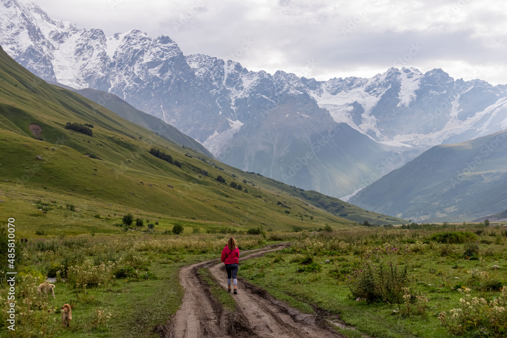 A female hiker on a hiking path leading to the Shkhara Glacier in the Greater Caucasus Mountain Range in Georgia, Svaneti Region, Ushguli. Snow-capped mountains in the back. Wanderlust. Wilderness.