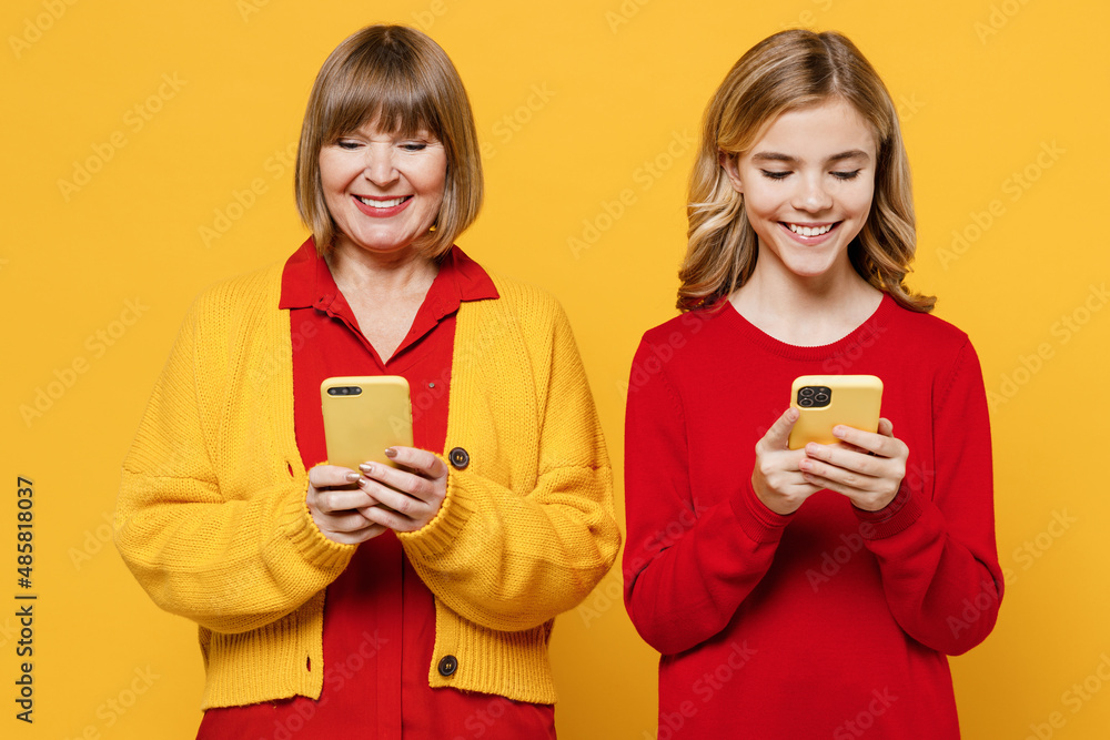 Happy woman 50s in red shirt have fun with teenager girl 12-13 years old. Grandmother granddaughter hold in hand use mobile cell phone isolated on plain yellow background. Family lifestyle concept Happy woman 50s in red shirt have fun with teenager girl 12-13 years old. Grandmother granddaughter hold in hand use mobile cell phone isolated on plain yellow background. Family lifestyle concept