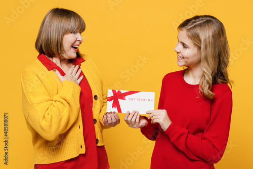 Happy thankful woman 50s in red shirt have fun with teenager girl 12-13 year old Grandmother granddaughter hold give gift certificate coupon voucher card for store isolated on plain yellow background
