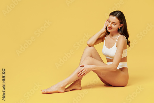 Full size young sexy brunette woman 20s wearing white underwear with perfect fit figure sitting on floor touch legs soft skin isolated on plain yellow color background. People female beauty concept