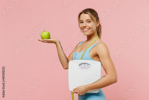 Side view happy young sporty fitness trainer instructor woman wear blue tracksuit spend time in home gym hold apple measure tape scales isolated on pastel plain pink background Workout sport concept