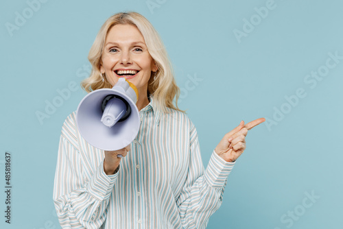 Elderly smiling woman 50s wear striped shirt hold scream in megaphone announces discounts sale Hurry up point finger aside on workspace isolated on plain pastel light blue color background studio