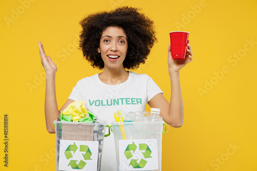 Young woman of African American ethnicity 20s in white volunteer t-shirt put plastic cup to boxes for waste sorting sprea hand isolated on plain yellow background. Voluntary free work help concept photo