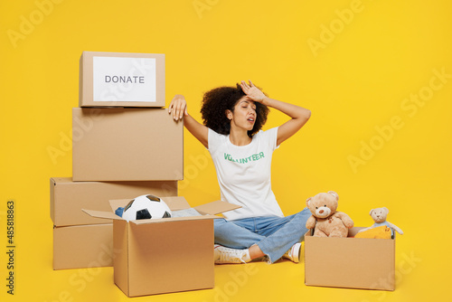 Full body tired young woman of African American ethnicity wear white volunteer t-shirt sit near boxes with presents isolated on plain yellow background. Voluntary free work assistance help concept photo