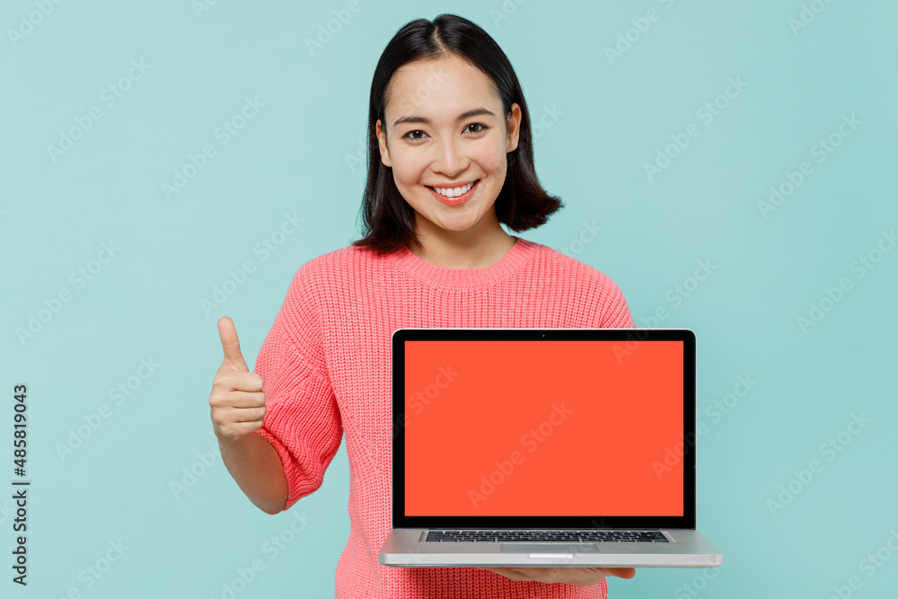 Young smiling happy woman of Asian ethnicity 20s wearing pink sweater hold use work on laptop pc computer with blank screen workspace area show thumb up isolated on pastel plain light blue background Young smiling happy woman of Asian ethnicity 20s wearing pink sweater hold use work on laptop pc computer with blank screen workspace area show thumb up isolated on pastel plain light blue background