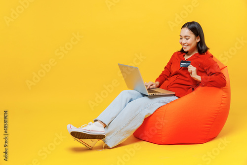 Full size young woman of Asian ethnicity 20s wear casual clothes sit in bag chair using laptop pc computer hold credit bank card do online shopping isolated on plain yellow background studio portrait photo