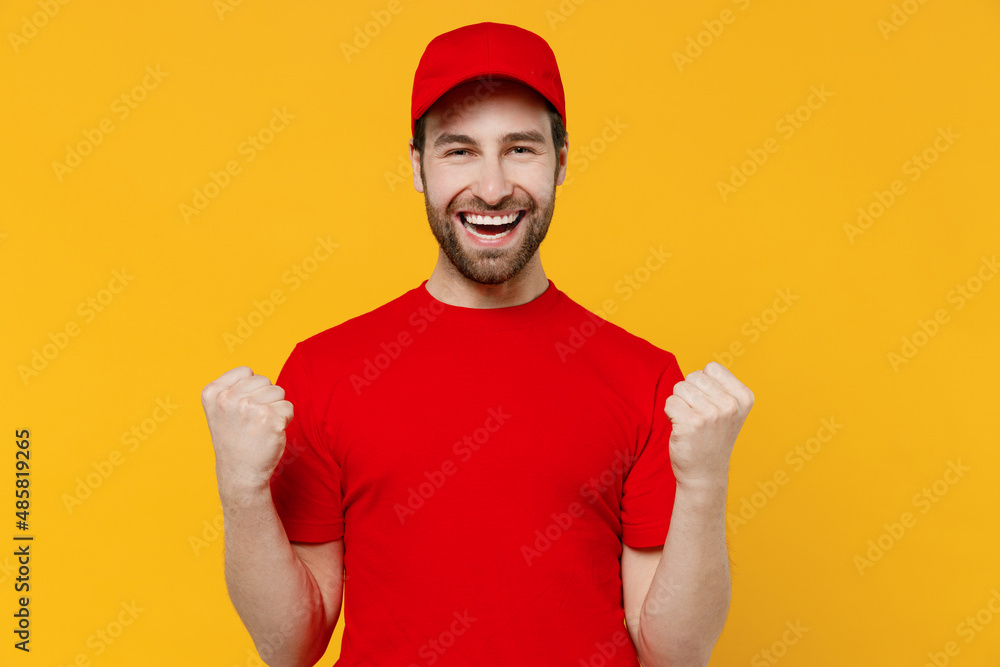 Naklejka premium Professional fun delivery guy employee man in red cap T-shirt uniform workwear work as dealer courier doing winner gesture celebrate clenching fists say yes isolated on plain yellow background studio.