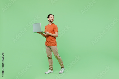 Full body young man 20s wear orange t-shirt hold use work on laptop pc computer look aside on workspace area isolated on plain pastel light green background studio portrait. People lifestyle concept. © ViDi Studio