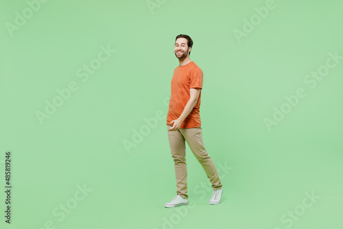 Full body side view smiling happy young man 20s wearing casual orange t-shirt walking going strolling isolated on plain pastel light green color background studio portrait. People lifestyle concept.
