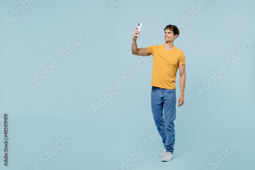 Full body young man 20s in yellow t-shirt doing selfie shot on mobile cell phone post photo on social network isolated on plain pastel light blue background studio portrait. People lifestyle concept