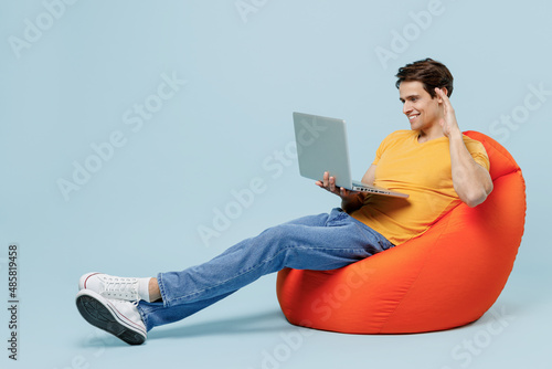 Full body side view fun young man 20s wearing yellow t-shirt sit in bag chair hold use work on laptop pc computer waving hand talk by video call isolated on plain pastel light blue background studio.