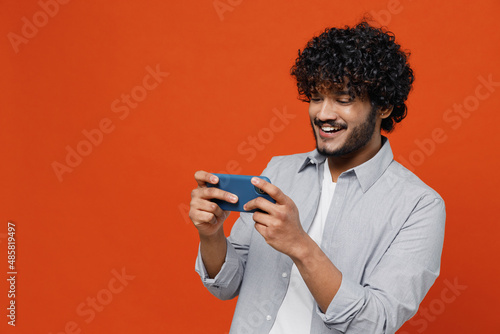 Gambling jubilant young bearded Indian man 20s wear blue shirt using play racing app on mobile cell phone hold gadget smartphone for pc video games isolated on plain orange background studio portrait