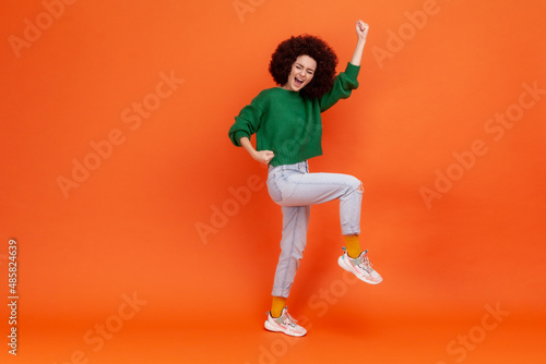Full length portrait of woman with Afro hairstyle wearing green casual style sweater screaming with happy expression, celebrating success. Indoor studio shot isolated on orange background.