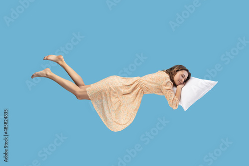 Relaxed girl in yellow dress levitating in mid-air, sleeping on stomach lying comfortable cozy on pillow, keeping eyes closed, watching peaceful dream. indoor studio shot isolated on blue