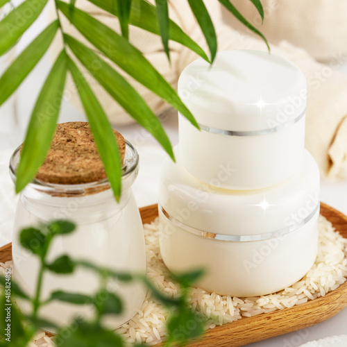 Fermented beauty care. Wellness composition with hande made rice water  creams and rice grain on a wooden plate on a white table with palm leaves. Natural homemade cosmetics based on fermented product