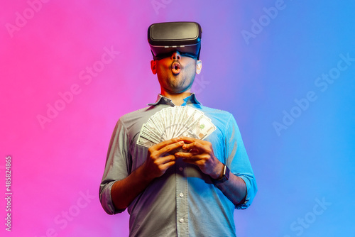 Portrait of amzed man in shirt to get money, holding dollars, illusion of rich millionaire, playing virtual reality game. Indoor studio shot isolated on colorful neon light background. photo