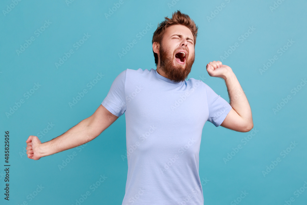 Portrait of sleepless handsome bearded man standing and yawning with closed eyes and raised arms, feels sleepy, needs more energy. Indoor studio shot isolated on blue background. Portrait of sleepless handsome bearded man standing and yawning with closed eyes and raised arms, feels sleepy, needs more energy. Indoor studio shot isolated on blue background.