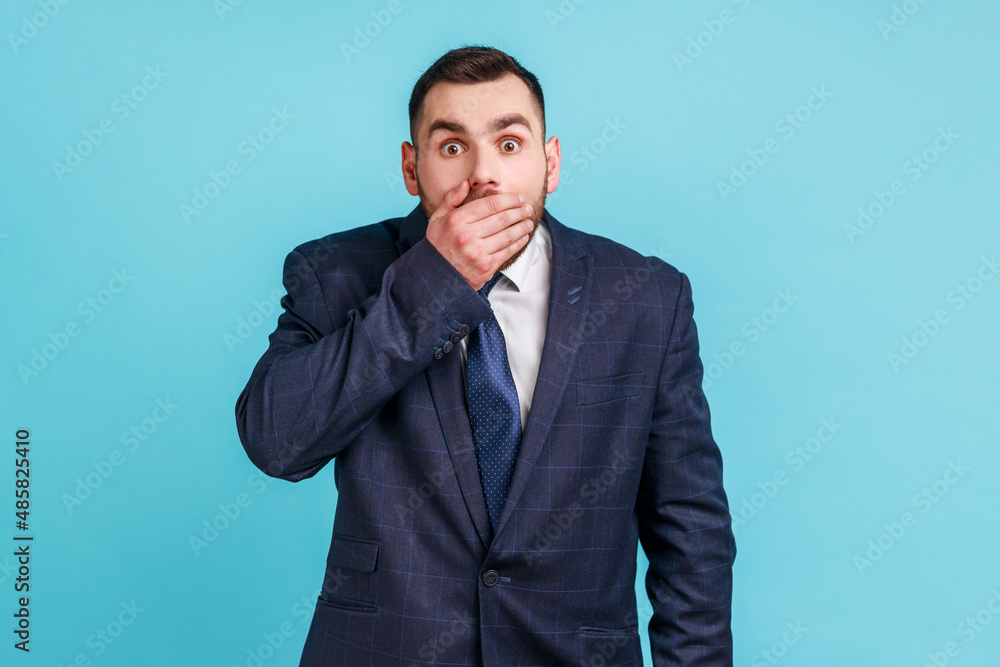 I won't tell. Portrait of amazed businessman wearing official style suit covering mouth with hand, keeping secret, terrified with shocking news. Indoor studio shot isolated on blue background. I won't tell. Portrait of amazed businessman wearing official style suit covering mouth with hand, keeping secret, terrified with shocking news. Indoor studio shot isolated on blue background.