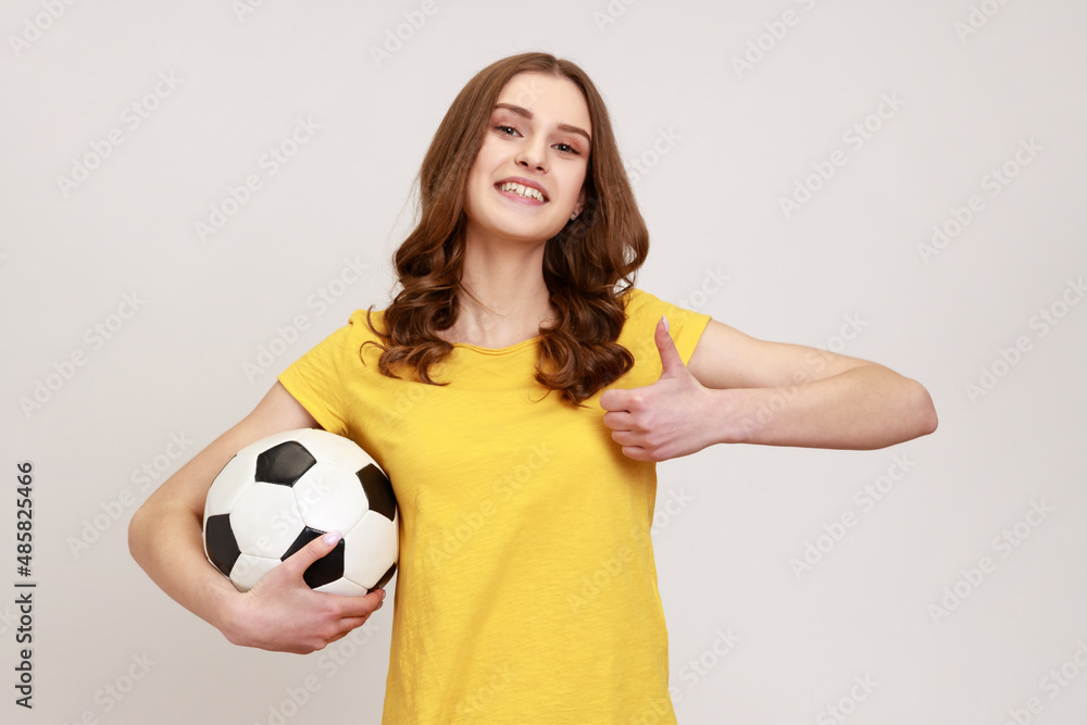 Young beautiful brown haired teenager girl holding soccer ball, looking at camera with toothy smile. showing thumb up, wearing casual attire. Indoor studio shot isolated on gray background. Young beautiful brown haired teenager girl holding soccer ball, looking at camera with toothy smile. showing thumb up, wearing casual attire. Indoor studio shot isolated on gray background.