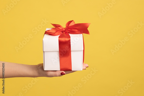 Closeup side view of woman hand holding out white gift box with red ribbon, giving present on holiday, bonuses and surprises concept. Indoor studio shot isolated on yellow background. photo