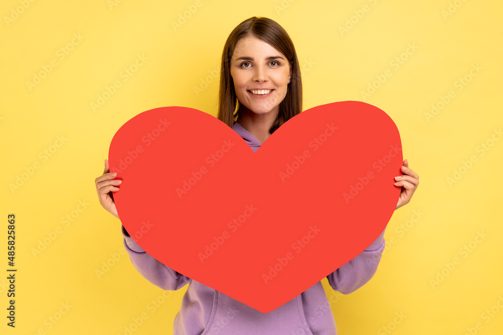 Portrait of happy cheerful woman holding in hands big red paper heart and smiling at camera, love and romance, wearing purple hoodie. Indoor studio shot isolated on yellow background. Portrait of happy cheerful woman holding in hands big red paper heart and smiling at camera, love and romance, wearing purple hoodie. Indoor studio shot isolated on yellow background.