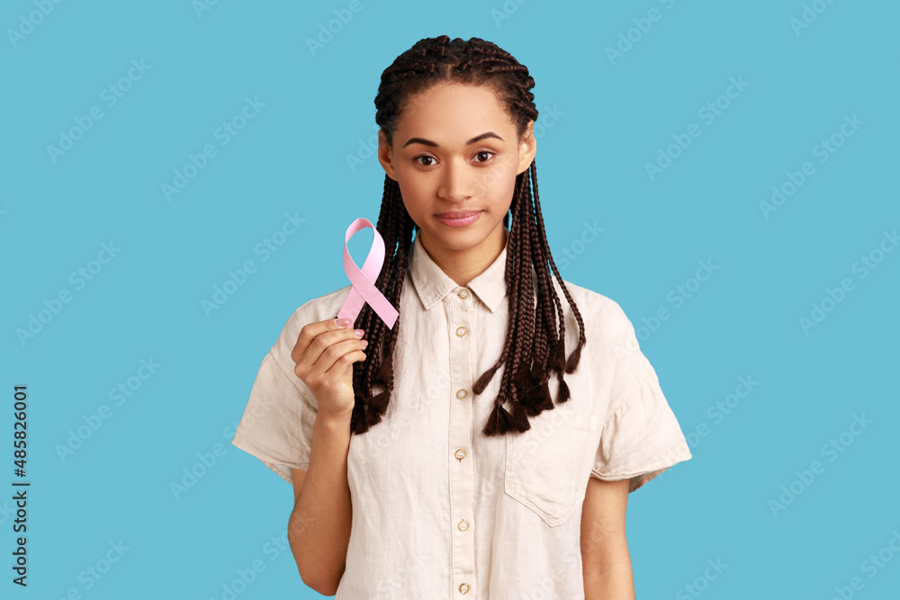 Portrait of attractive positive woman with black dreadlocks holding pink ribbon, symbol of breast cancer awareness, wearing white shirt. Indoor studio shot isolated on blue background.
