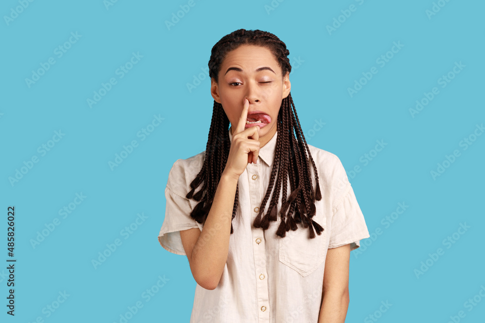 Portrait of funny impolite woman with black dreadlocks making crazy face with tongue out and picking her nose, bad manners, wearing white shirt. Indoor studio shot isolated on blue background. Portrait of funny impolite woman with black dreadlocks making crazy face with tongue out and picking her nose, bad manners, wearing white shirt. Indoor studio shot isolated on blue background.