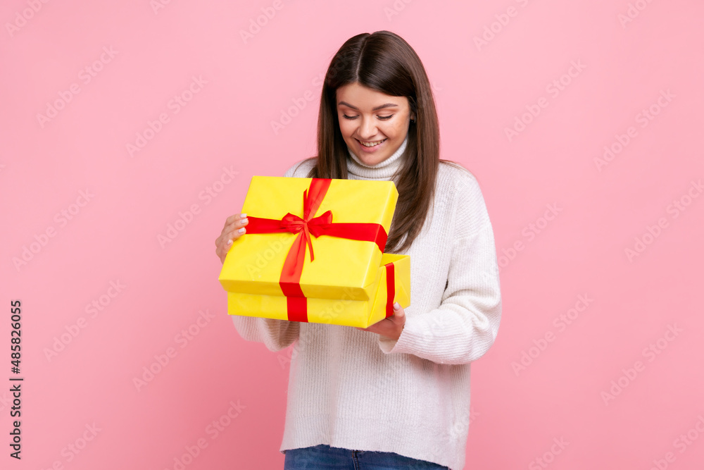 Positive happy dark haired girl looking inside wrapped present box, open gift, expressing positive, wearing white casual style sweater. Indoor studio shot isolated on pink background. Positive happy dark haired girl looking inside wrapped present box, open gift, expressing positive, wearing white casual style sweater. Indoor studio shot isolated on pink background.