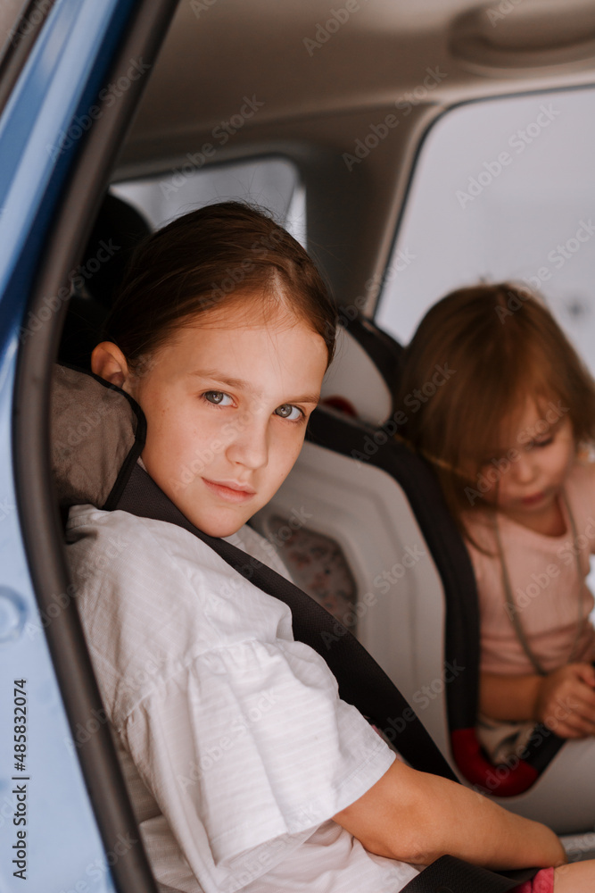 Little girl sitting in opened car and fastening seat belt with soft grey pillow. Safety concept.