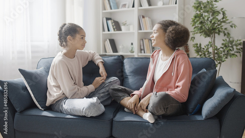 Happy teenage sisters talking, sitting on the sofa at home, communication