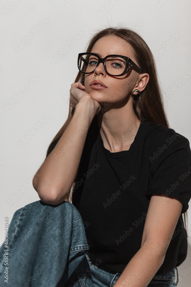 Pretty glamour young woman with stylish glasses eyewear in fashion black  mock-up t-shirt and jeans poses and sits in studio on a white background.  Sexy girl in trendy casual clothes Stock Photo