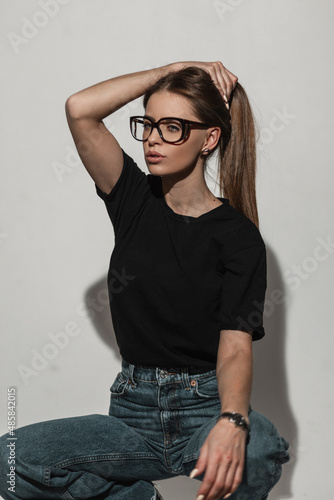 Cool pretty woman model with fashion glasses in black t-shirt mock-up and vintage jeans sits and poses in studio near white wall. Glamour sexy girl model in trendy clothes