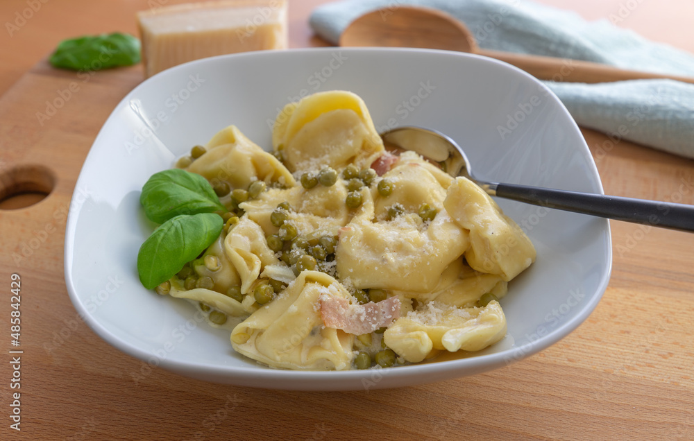 Tortellini alla panna, Tortellini with parmesan, basil, pea and boiled ham in a white plate with a spoon, focus on foreground Tortellini alla panna, Tortellini with parmesan, basil, pea and boiled ham in a white plate with a spoon, focus on foreground